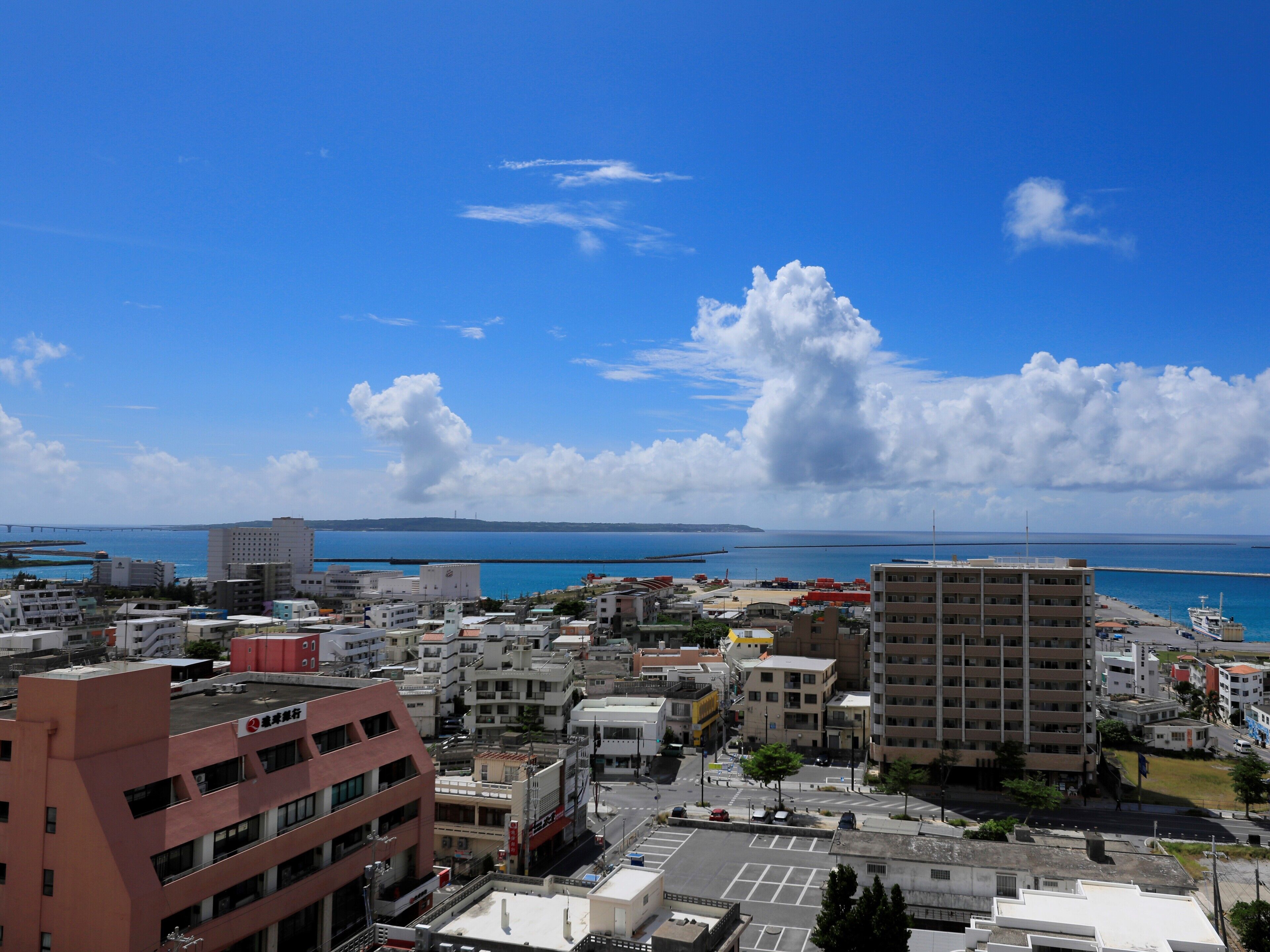 A coastal cityscape with a clear blue sky, a large body of water, and a variety of buildings including a prominent red building and several multi-story structures.
