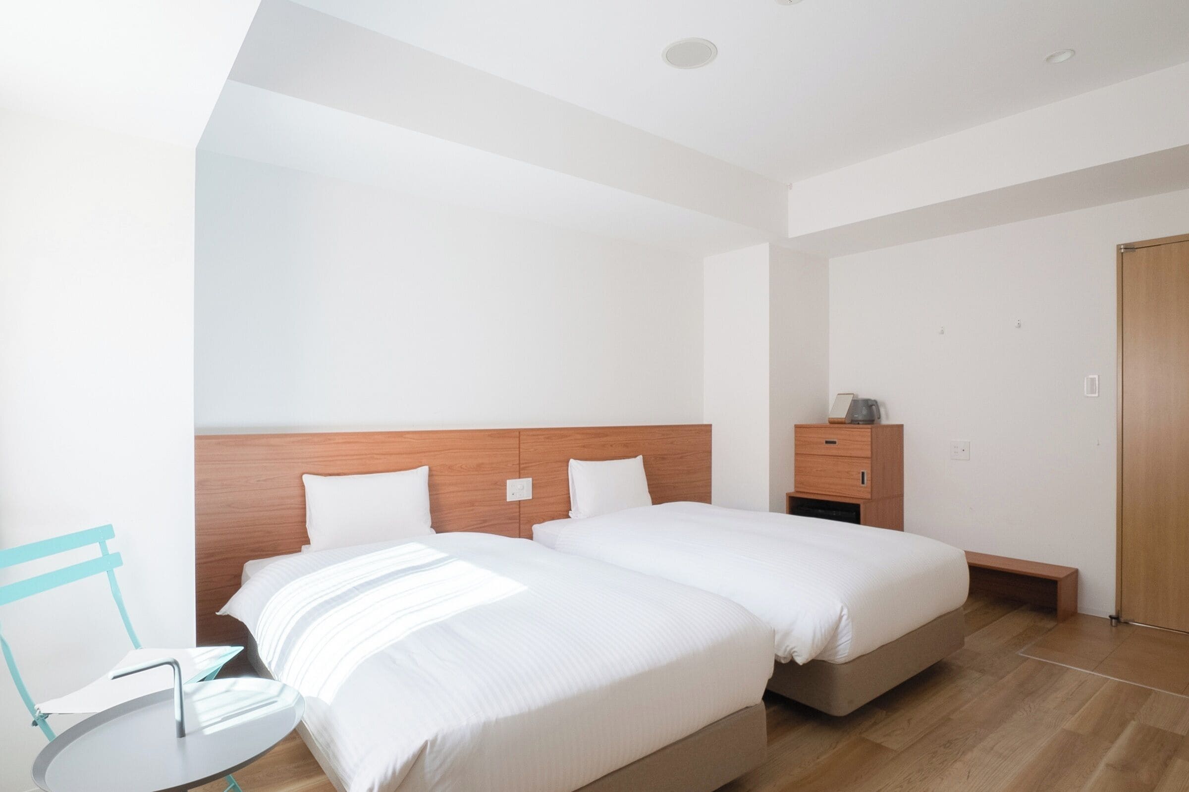 Quarto de hotel moderno com duas camas, cabeceira de madeira, mesa de cabeceira pequena e uma cadeira.