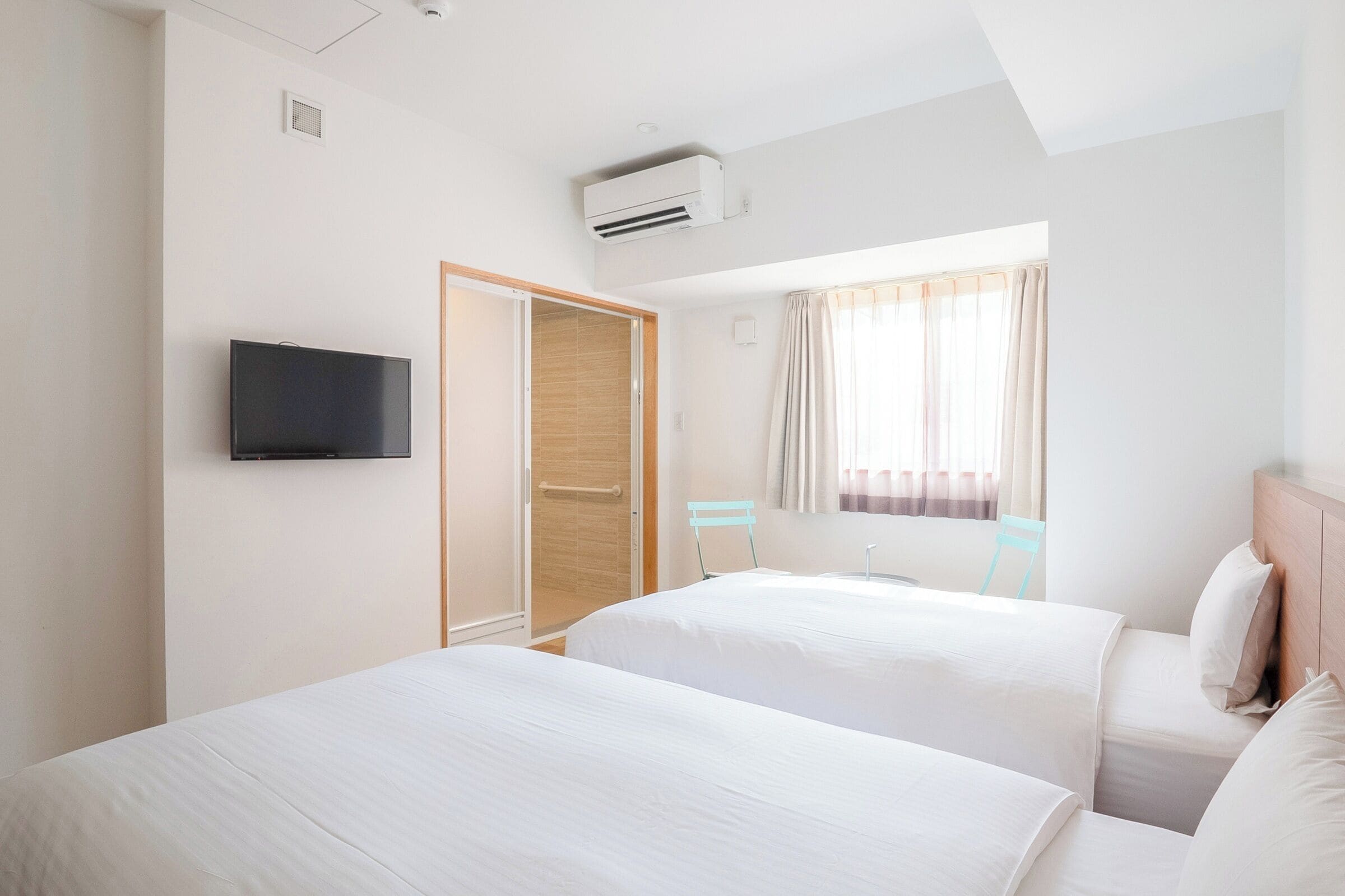 Ein Hotelzimmer mit zwei Betten, einem Fernseher, einer Klimaanlage, einem Fenster mit Vorhängen und einer Holztür.