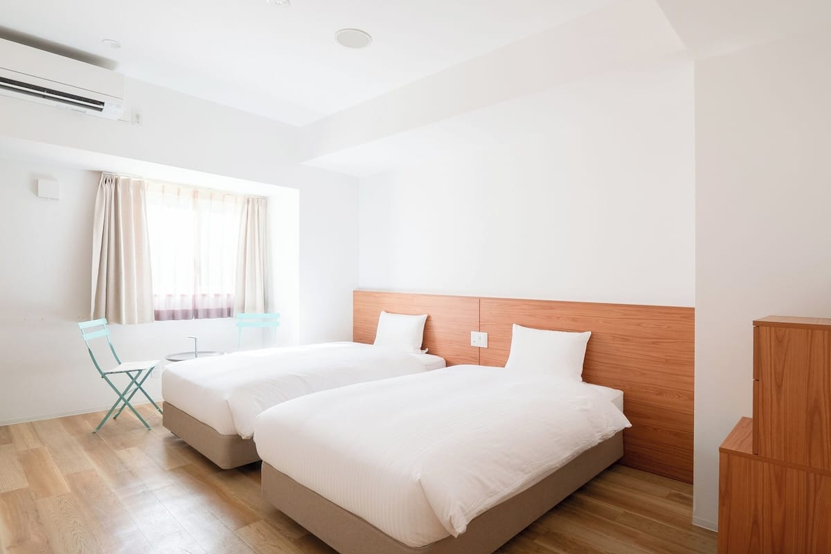 Une chambre d’hôtel avec deux lits, une tête de lit en bois, une fenêtre avec des rideaux, une petite table et une chaise.