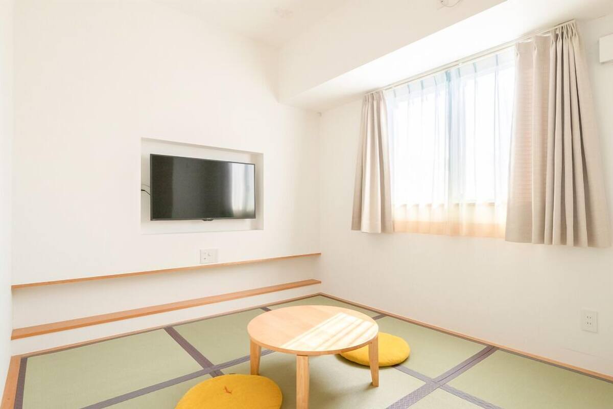 Une pièce avec un téléviseur fixé au mur, une fenêtre avec des rideaux, des tatamis au sol et une petite table en bois.
