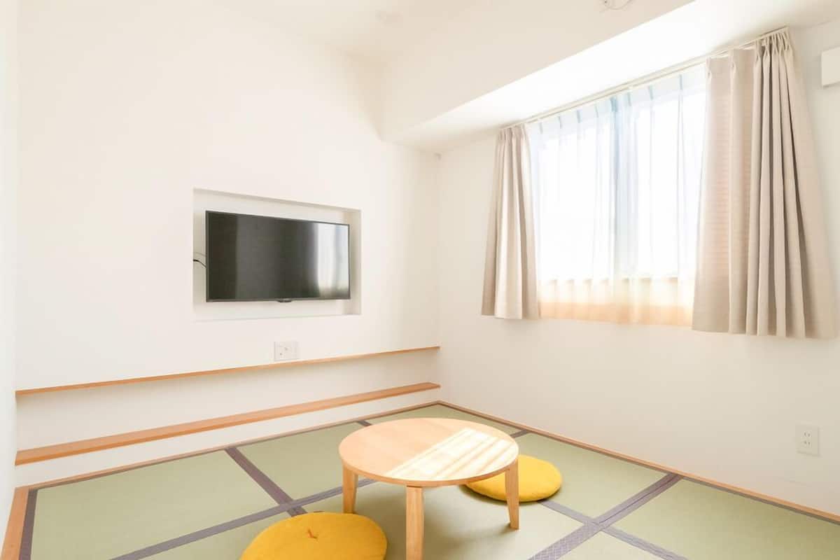 A room with a television mounted on the wall, a window with curtains, tatami mats on the floor, and a small wooden table.