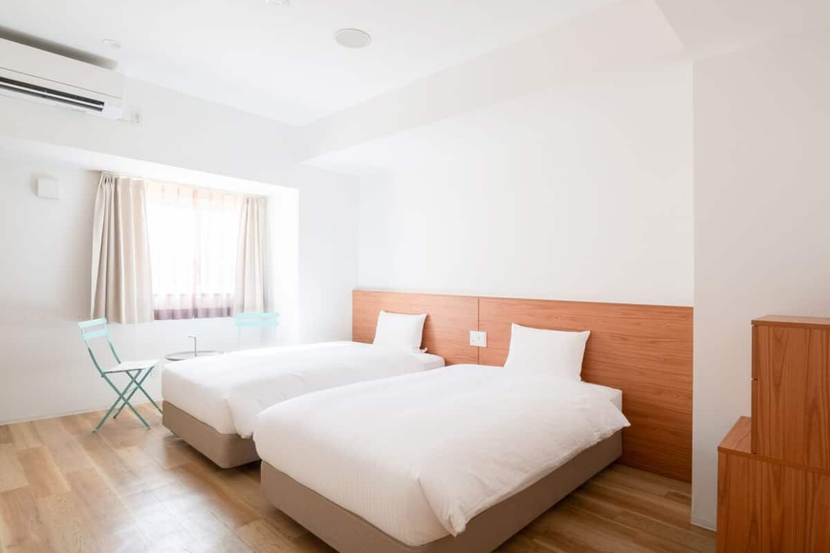 Une chambre d’hôtel avec deux lits, une tête de lit en bois, une fenêtre avec des rideaux, une chaise et une armoire en bois.