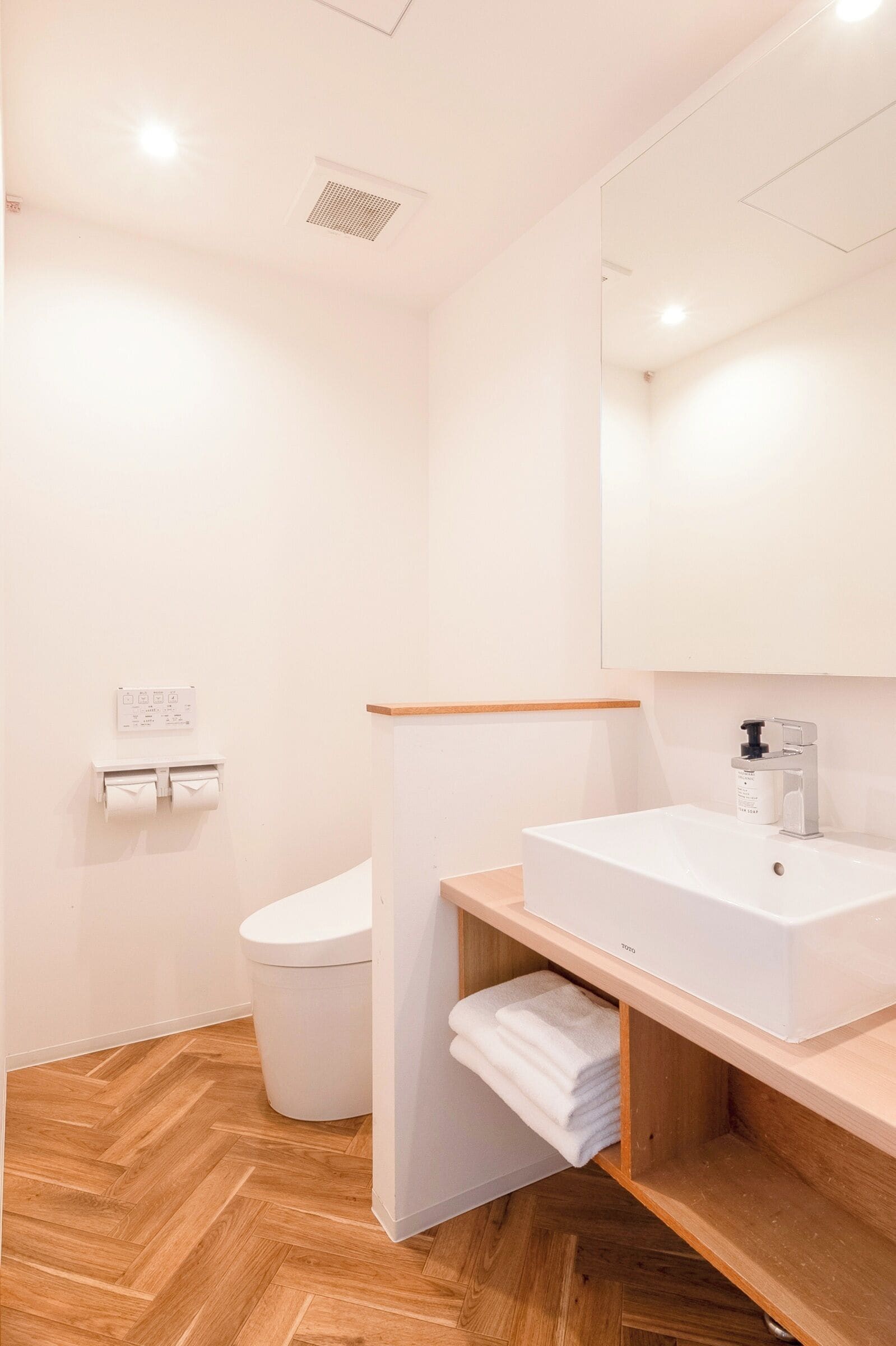Un baño moderno con lavamanos, espejo y váter blancos. Hay un estante de madera con toallas y un cubo de basura blanco.