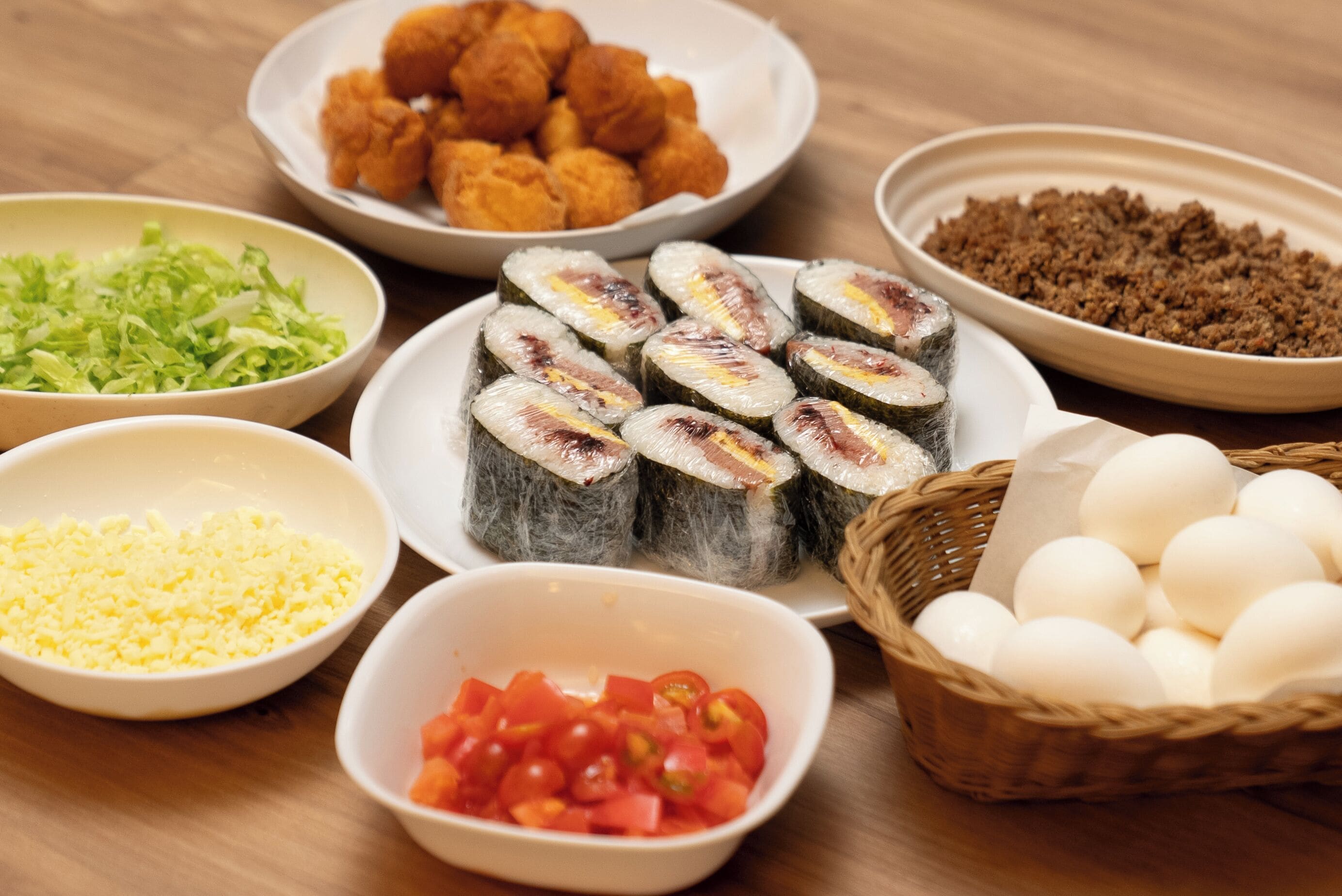Uma mesa com diversos alimentos, incluindo sushi, frango frito, alface desfiada, tomate picado e uma cesta de ovos.