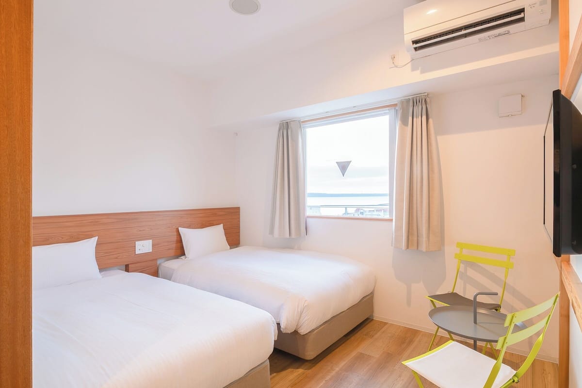 Une chambre d’hôtel avec deux lits, une fenêtre avec des rideaux, une petite table avec une chaise et un téléviseur à écran plat fixé au mur.