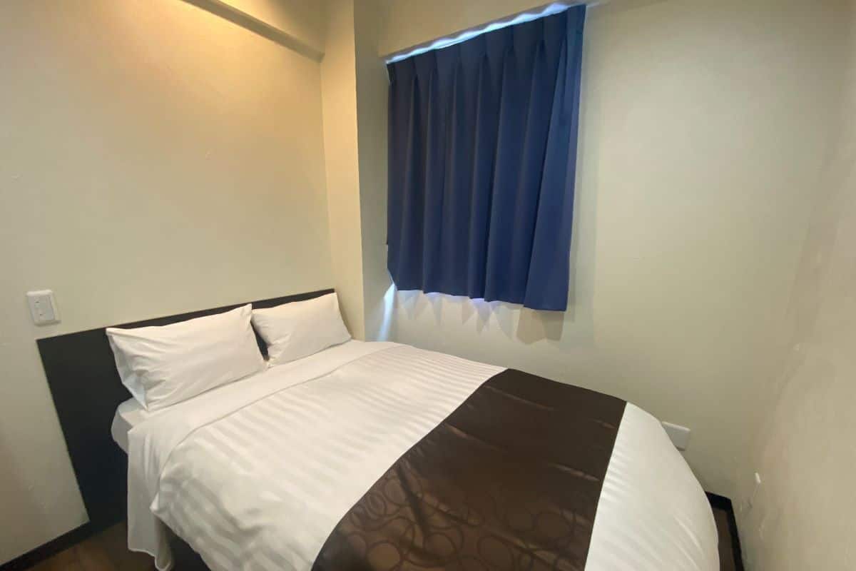 A hotel room with a bed, a headboard, a window with blue curtains, and a wall-mounted light.