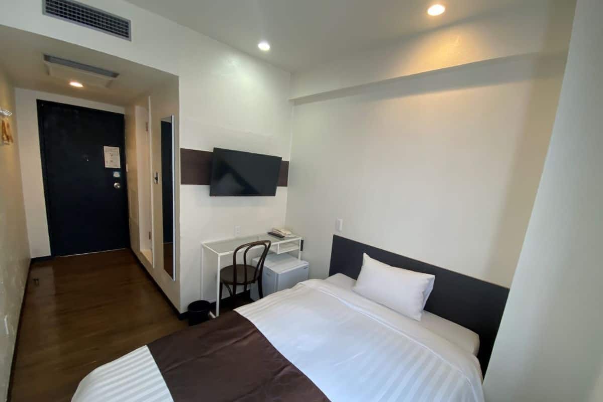 A hotel room with a bed, a desk, a chair, a TV mounted on the wall, and a door.