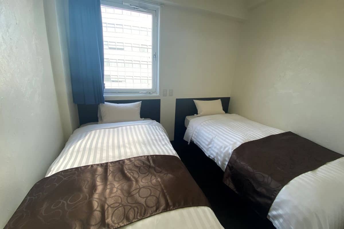 A room with two single beds, a window with curtains, and a plain wall.