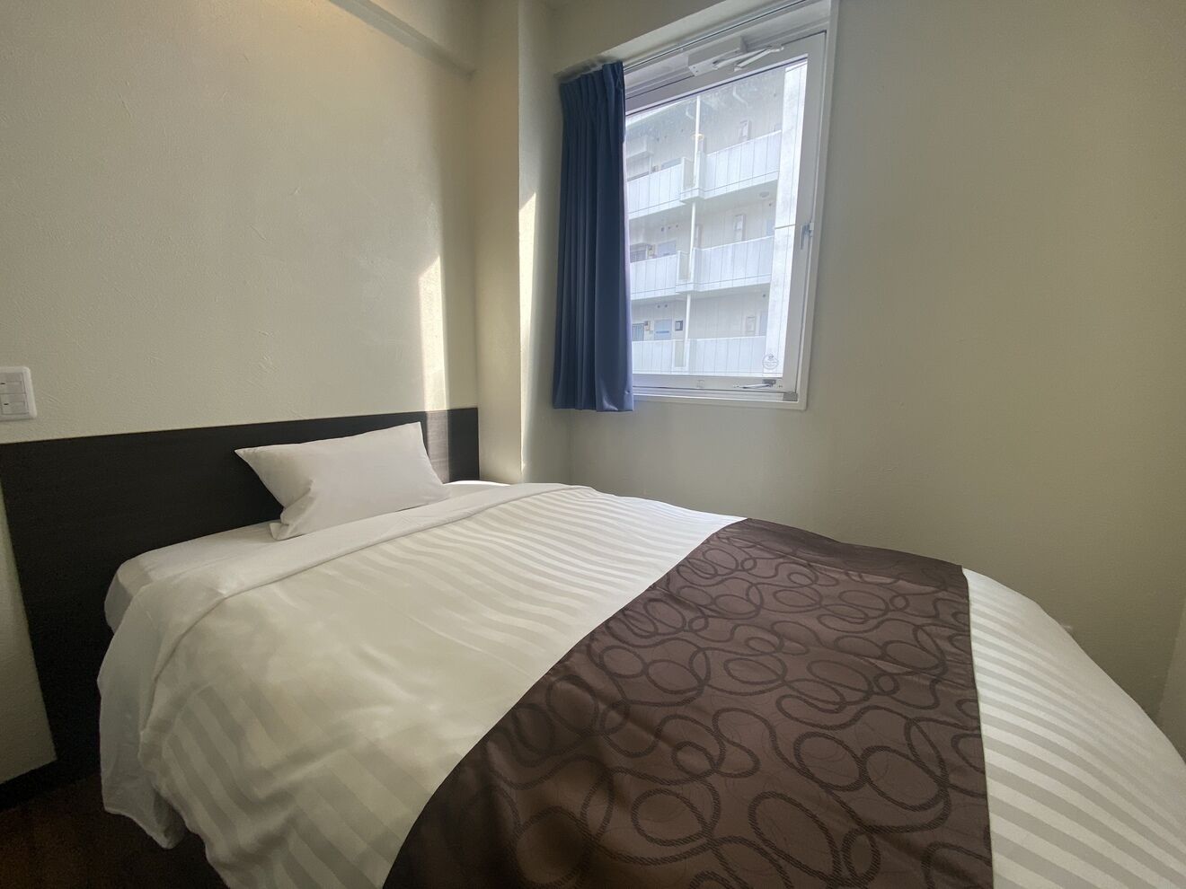 A hotel room with a bed, a window with curtains, and a view of buildings outside.