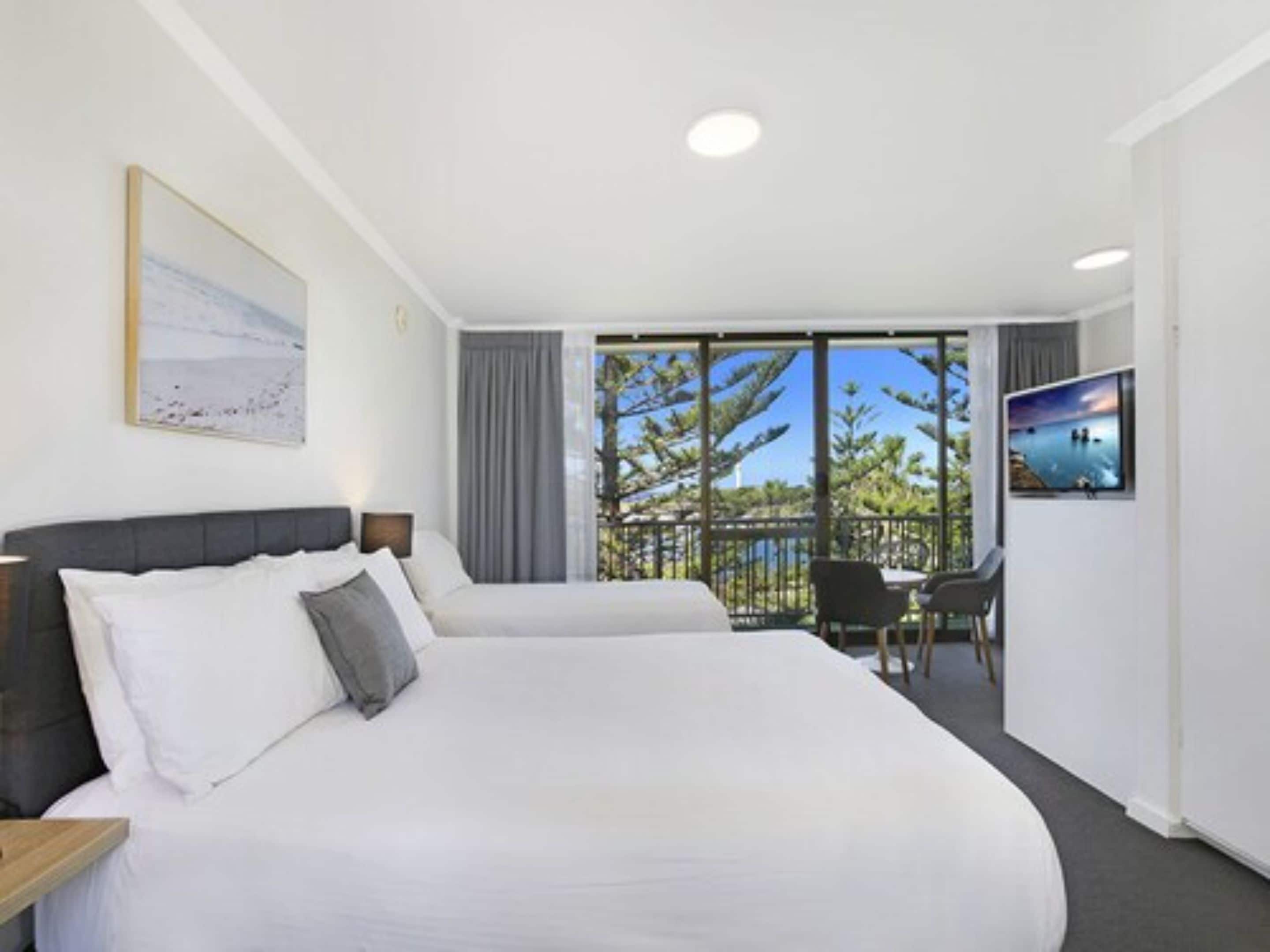 A hotel room with two beds, a balcony with a view of trees and a body of water, and a television on the wall.