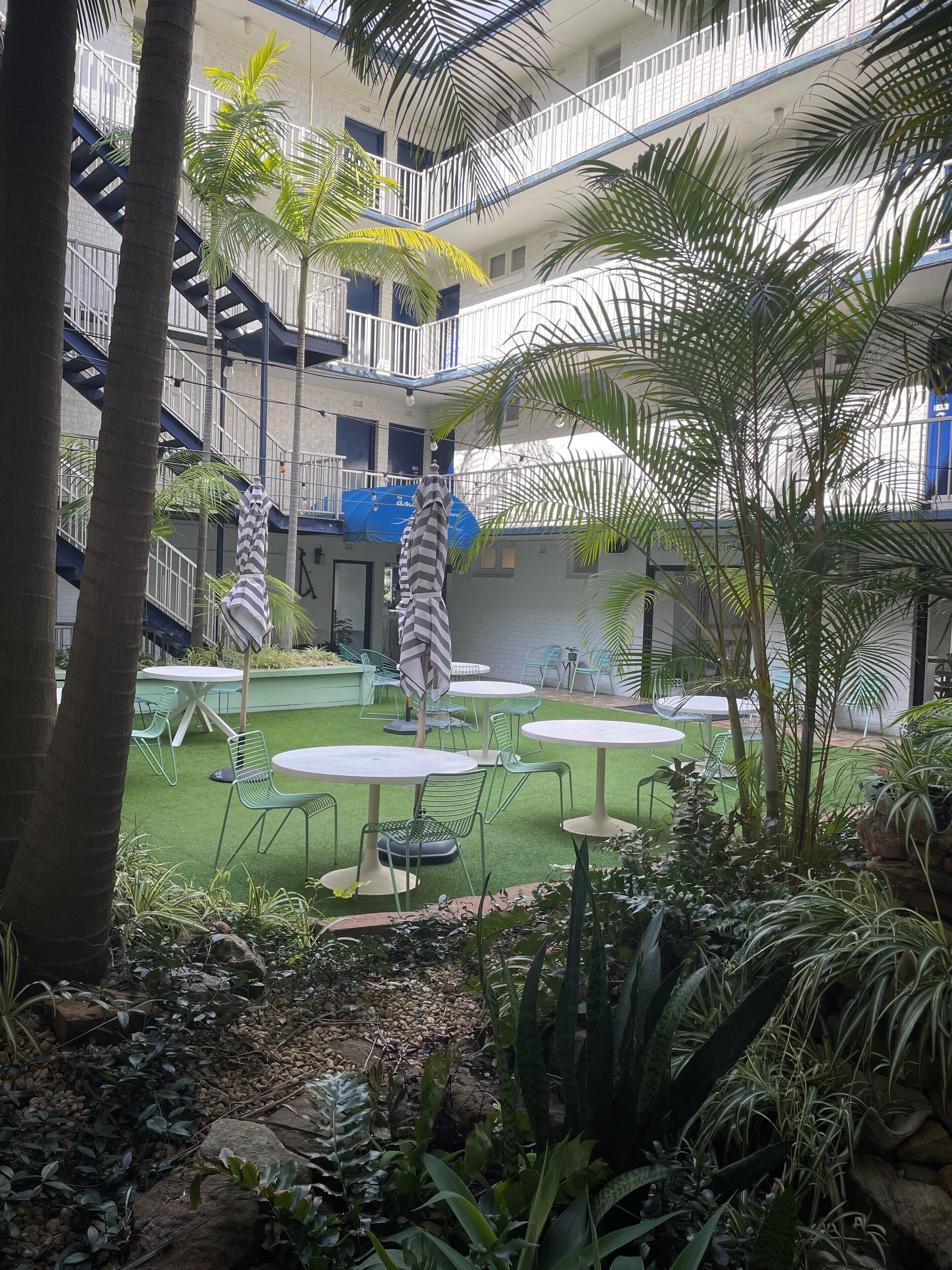 Un patio con mobiliario exterior, palmeras y un edificio de varios pisos con balcones.