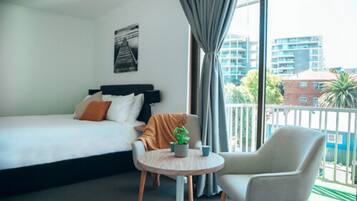 A hotel room with a bed, a small round table with a plant, a chair, and a view of buildings through large windows.