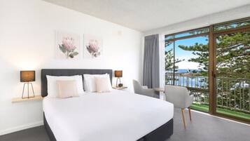 A modern hotel room with a large bed, a desk, and a chair. There are two wall-mounted lamps and a large window with a view of the outdoors.
