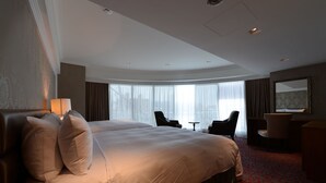 Habitación de hotel con una cama grande, un escritorio, una silla y vistas de la ciudad a través de amplias ventanas.