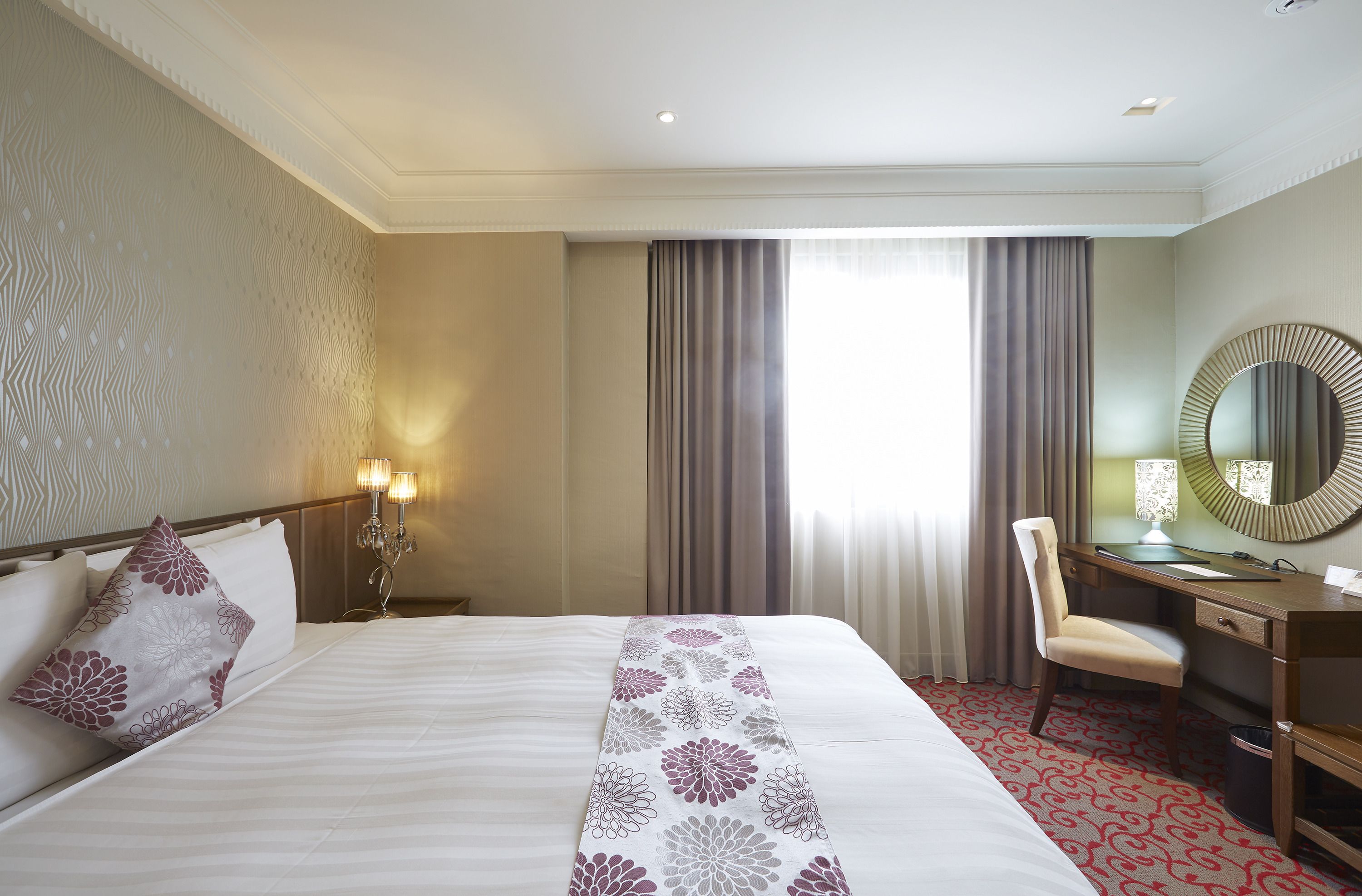 A hotel room with a large bed, a desk, a chair, a mirror, and a window with curtains.