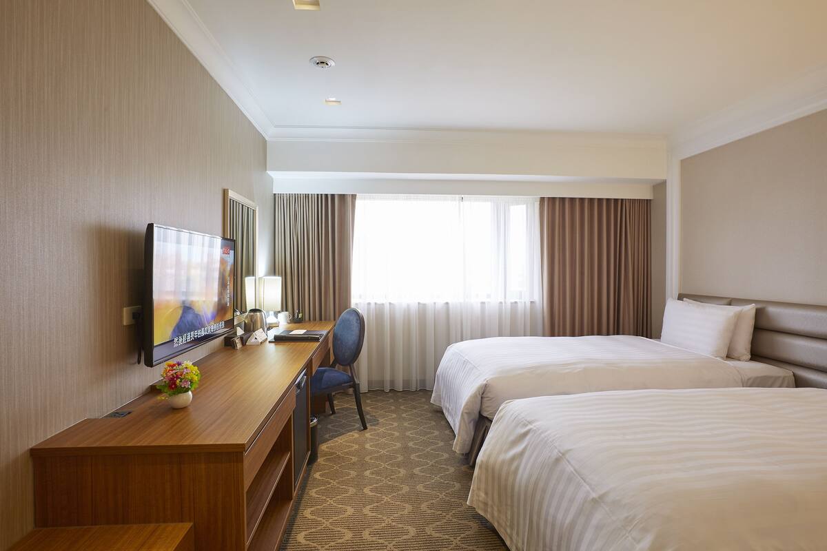 A hotel room with two beds, a desk with a chair, a television, and a window with curtains.
