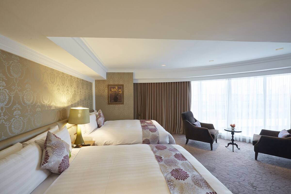 A hotel room with two beds, a sitting area with a table, and a large window with curtains.
