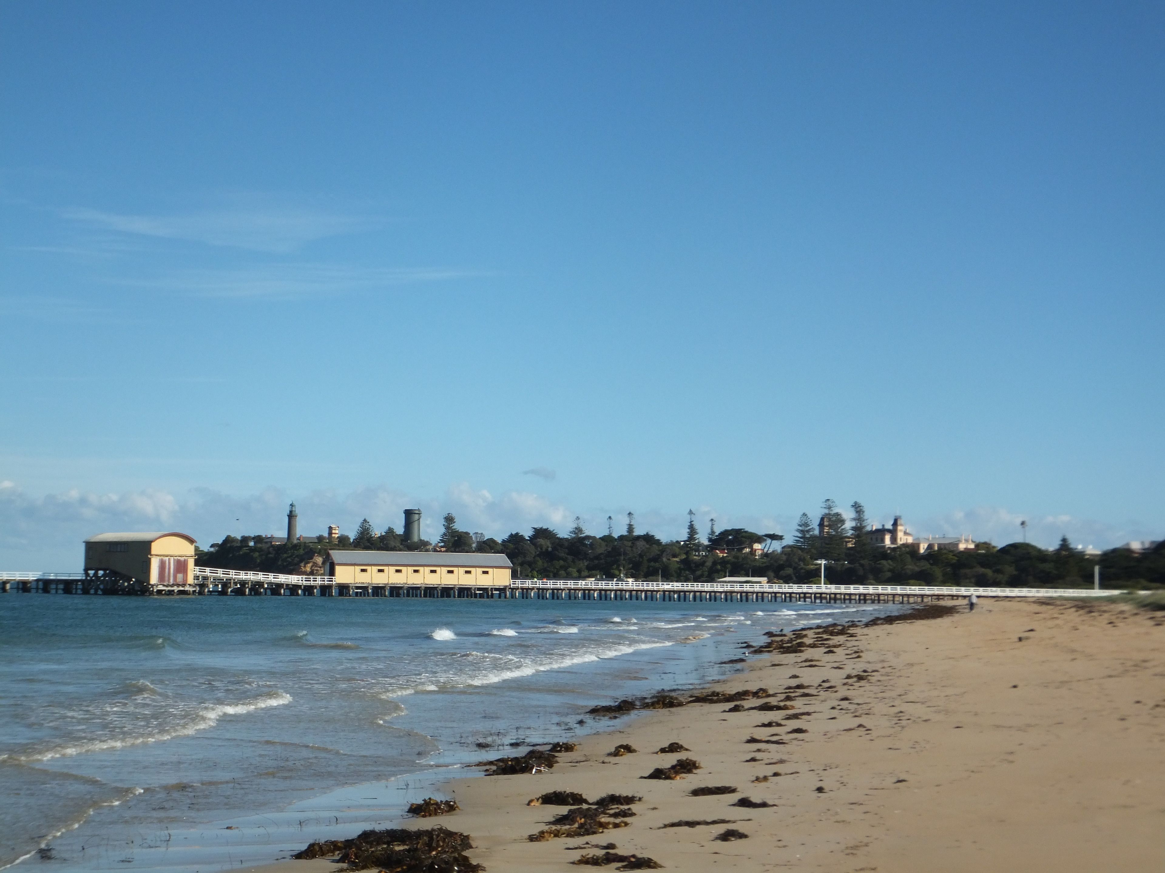 A beach with a pier extending into the water, featuring a yellow building and a white fence.