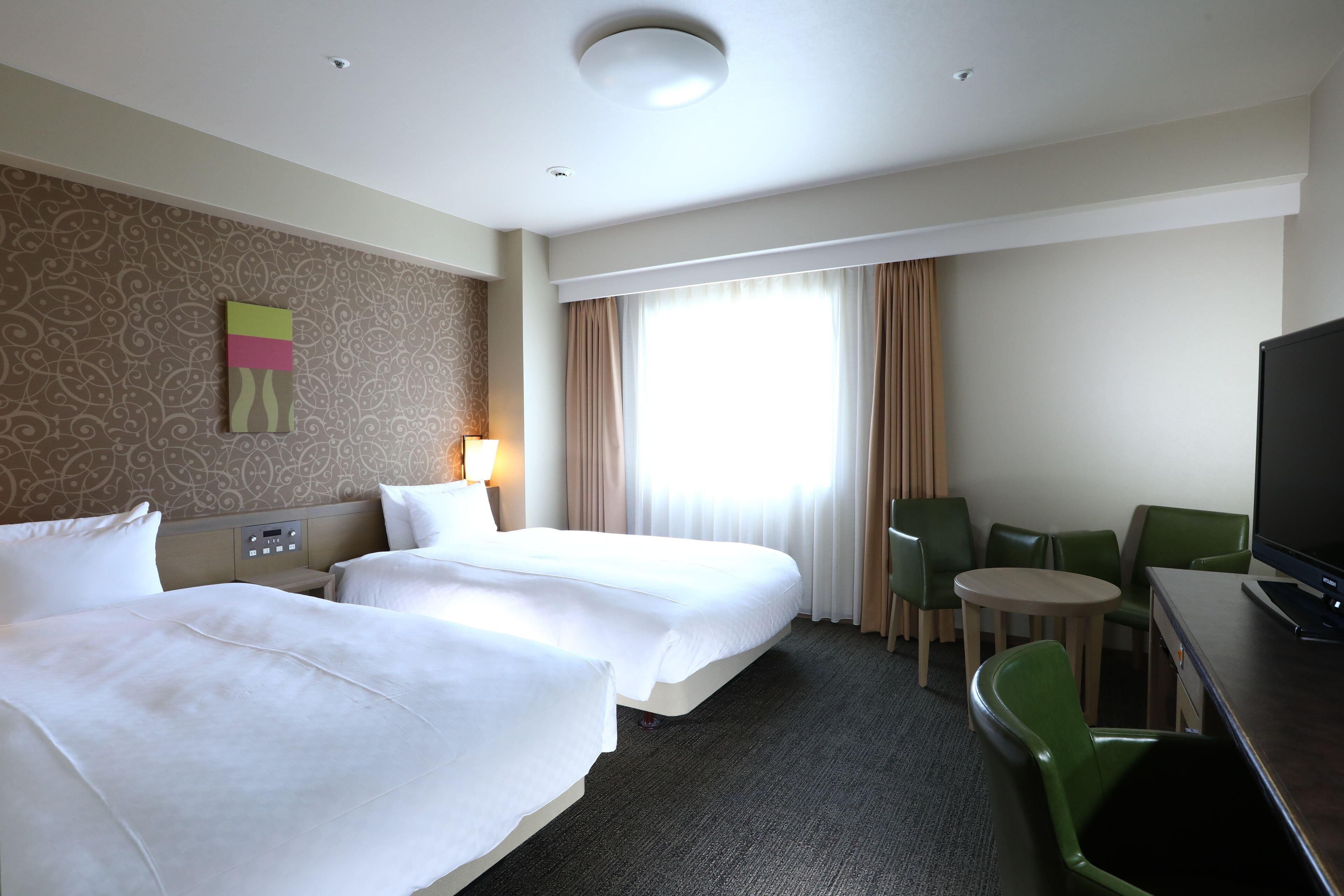 A hotel room with two beds, a desk with a TV, a small round table, and green chairs.