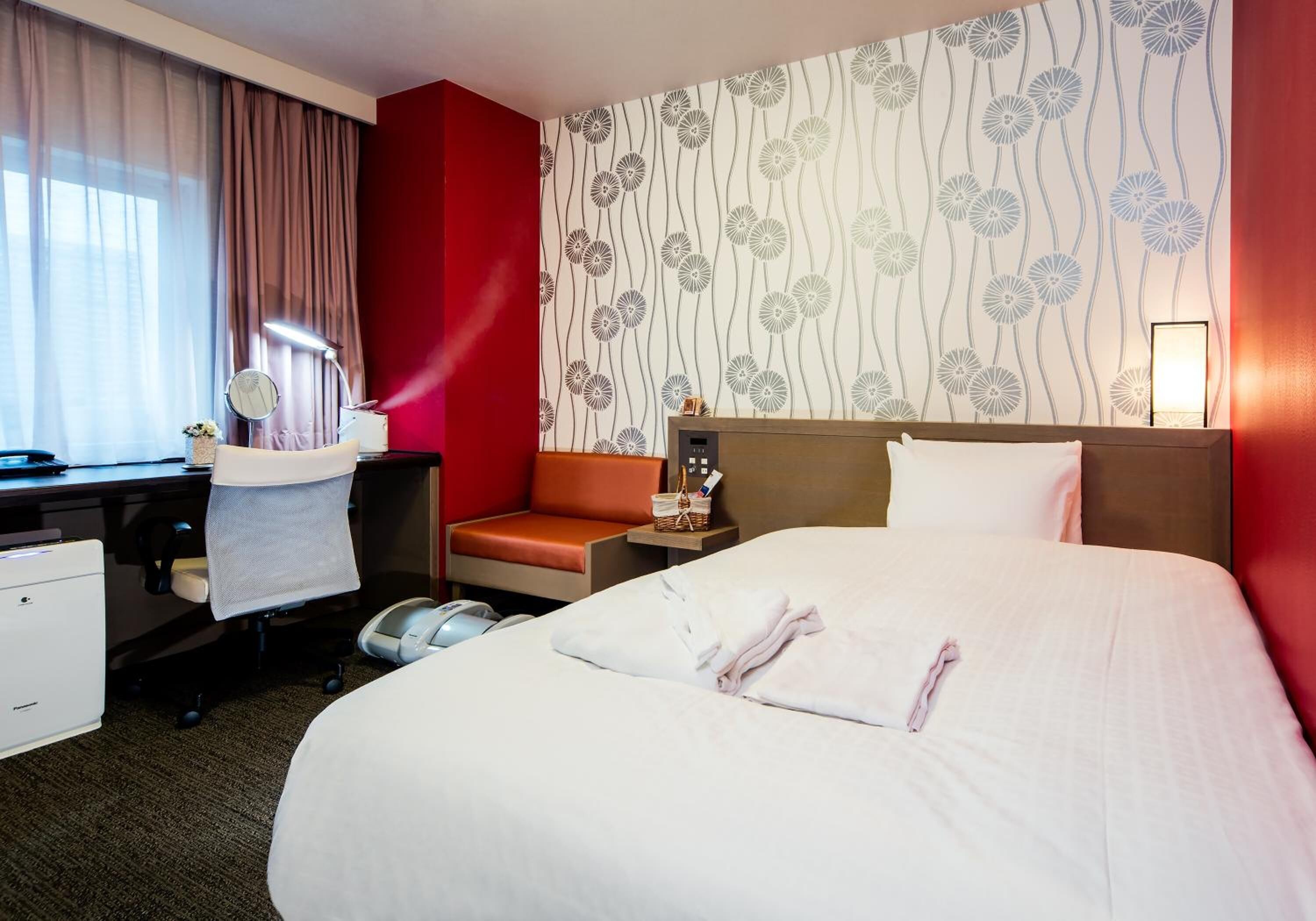 Une chambre d’hôtel avec un grand lit, un bureau avec un ordinateur, une chaise, une petite table avec une lampe et un mur d’accent de couleur rouge.