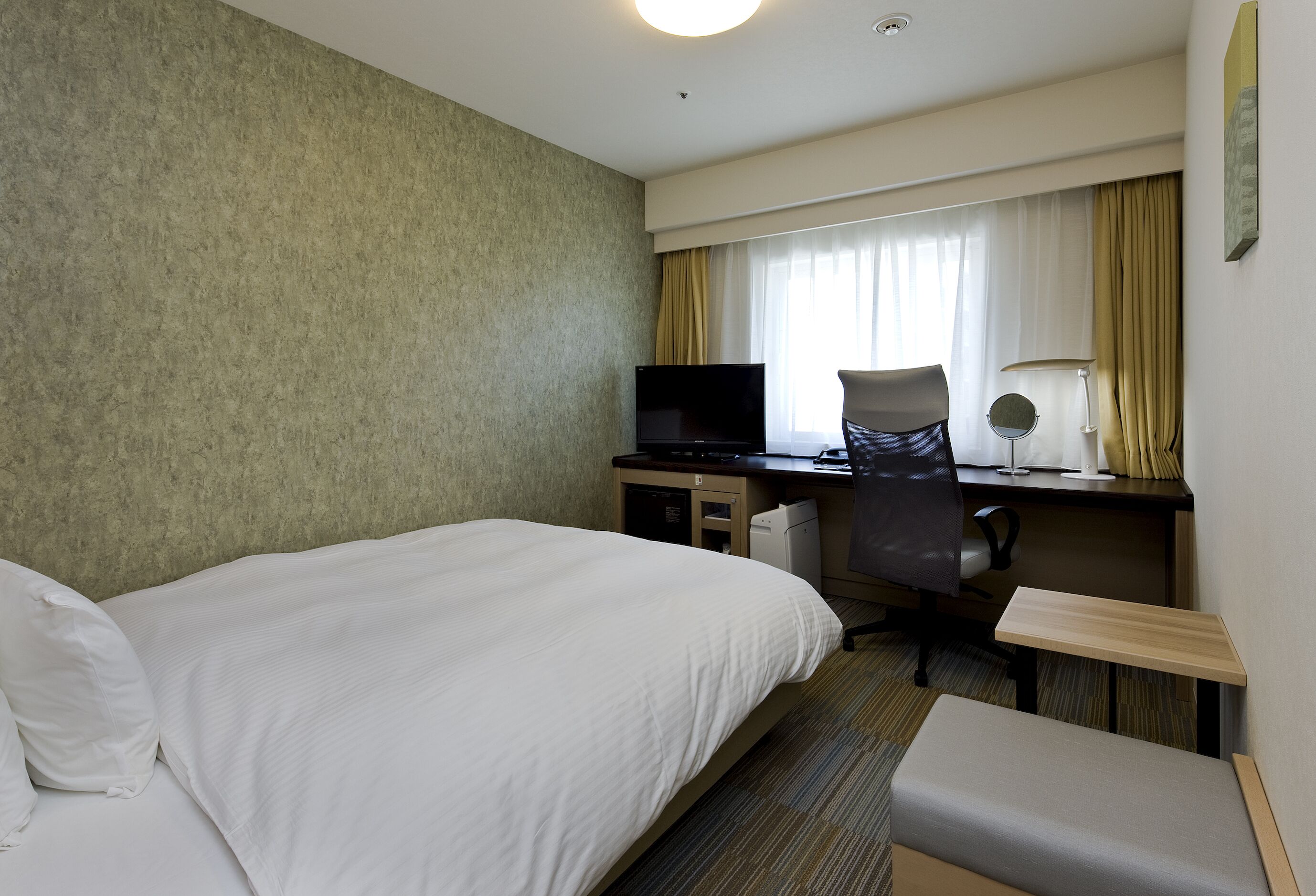 A hotel room with a bed, a desk with a computer, a chair, a lamp, and a small table.