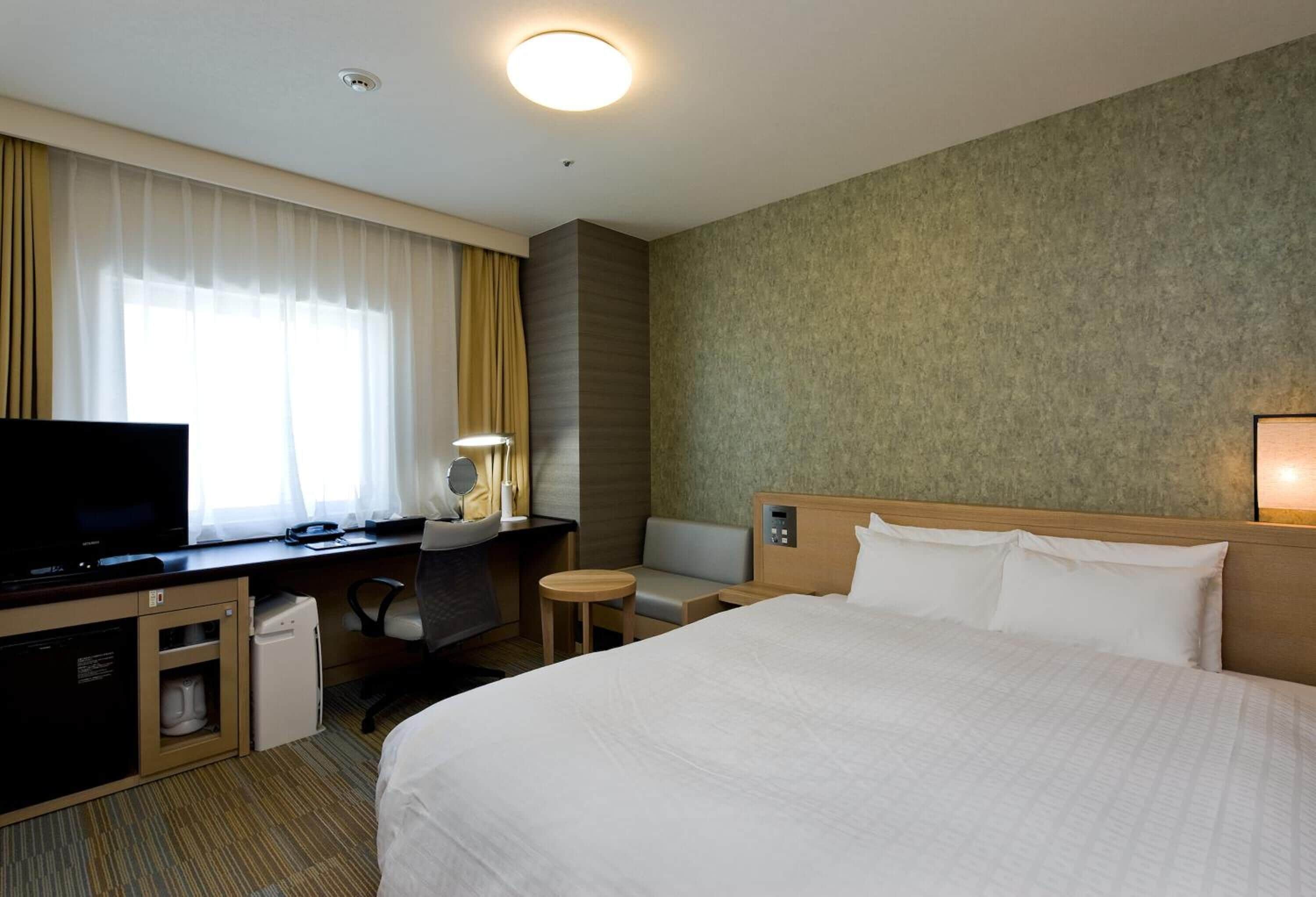 A hotel room with a bed, a desk with a television, a small table, and a chair.
