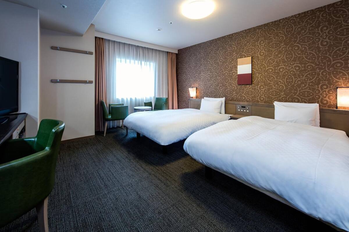 Ein Hotelzimmer mit zwei Einzelbetten, einem Schreibtisch mit Stuhl, einem Fernseher und einer Wand mit Muster.