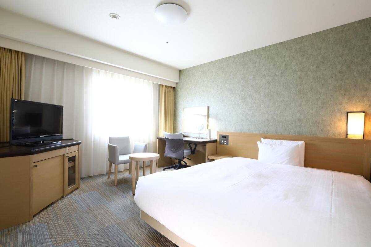 Ein Hotelzimmer mit einem großen Bett, einem Schreibtisch mit Stuhl, einem Fernseher und einem Fenster mit Vorhängen.