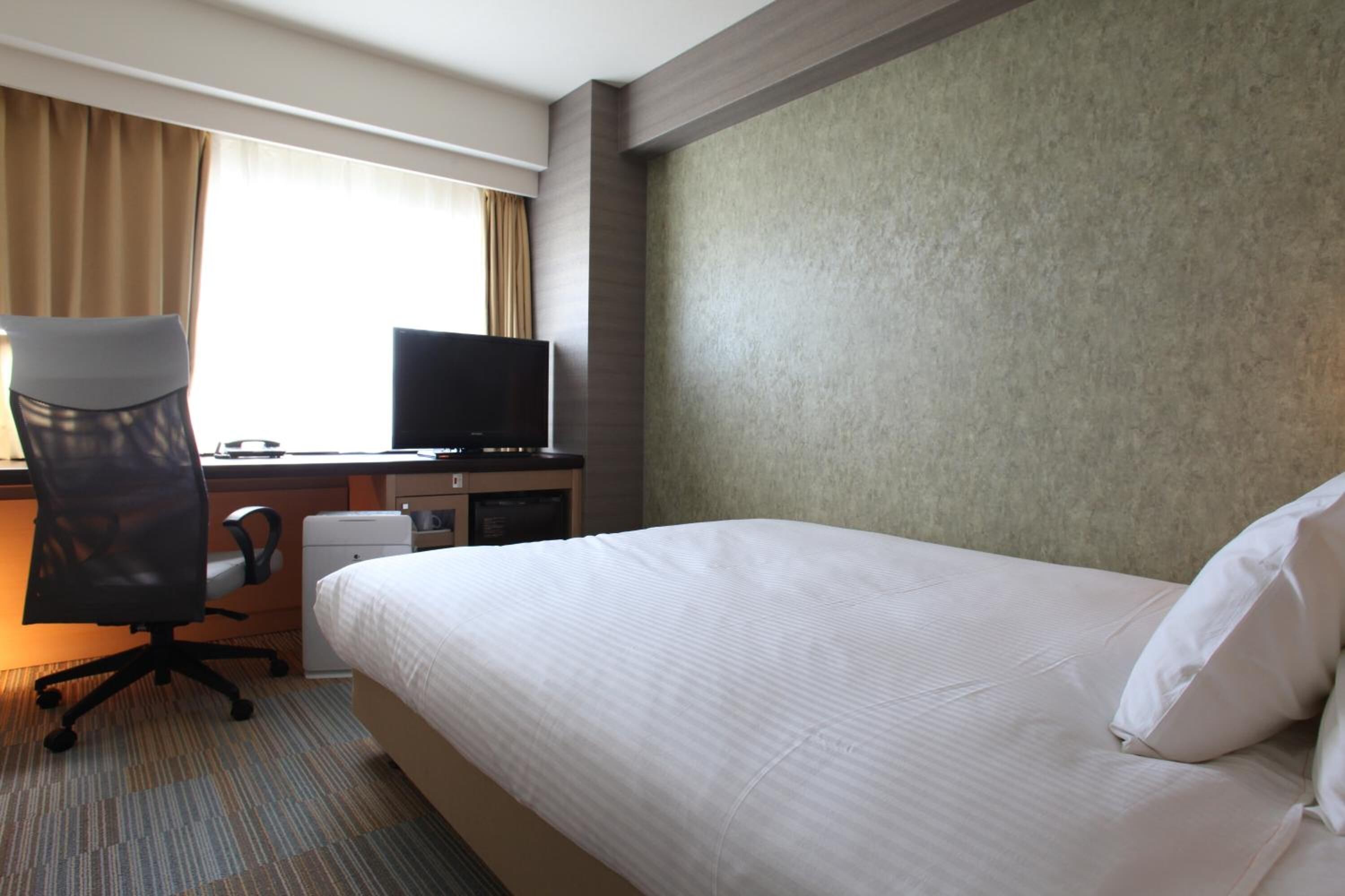 Una habitación de hotel con una cama grande, un escritorio con computadora y televisor, y un ventanal con cortinas.