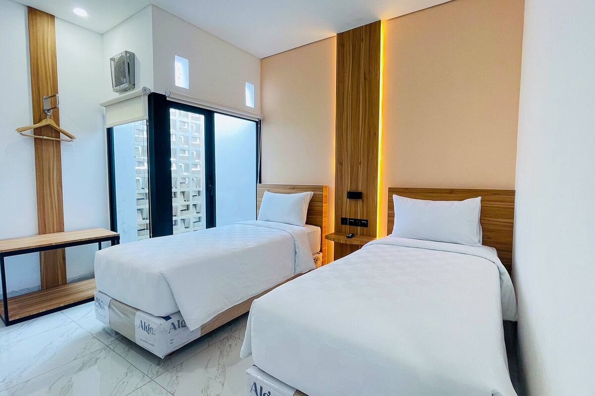 Ein Hotelzimmer mit zwei Betten, einem Holzschrank, einer Bank und einem großen Fenster mit Jalousien.