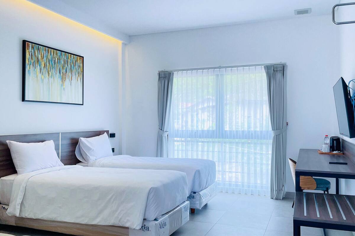 Ein Hotelzimmer mit zwei Betten, einem Schreibtisch, einem Stuhl und einem großen Fenster mit Vorhängen.