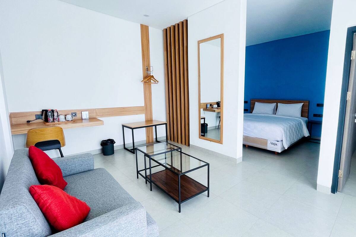 Ein modernes Hotelzimmer mit grauem Sofa, einer Holzkommode, einem Glastisch, einem Bett mit weißer Bettwäsche und einem großen Spiegel.