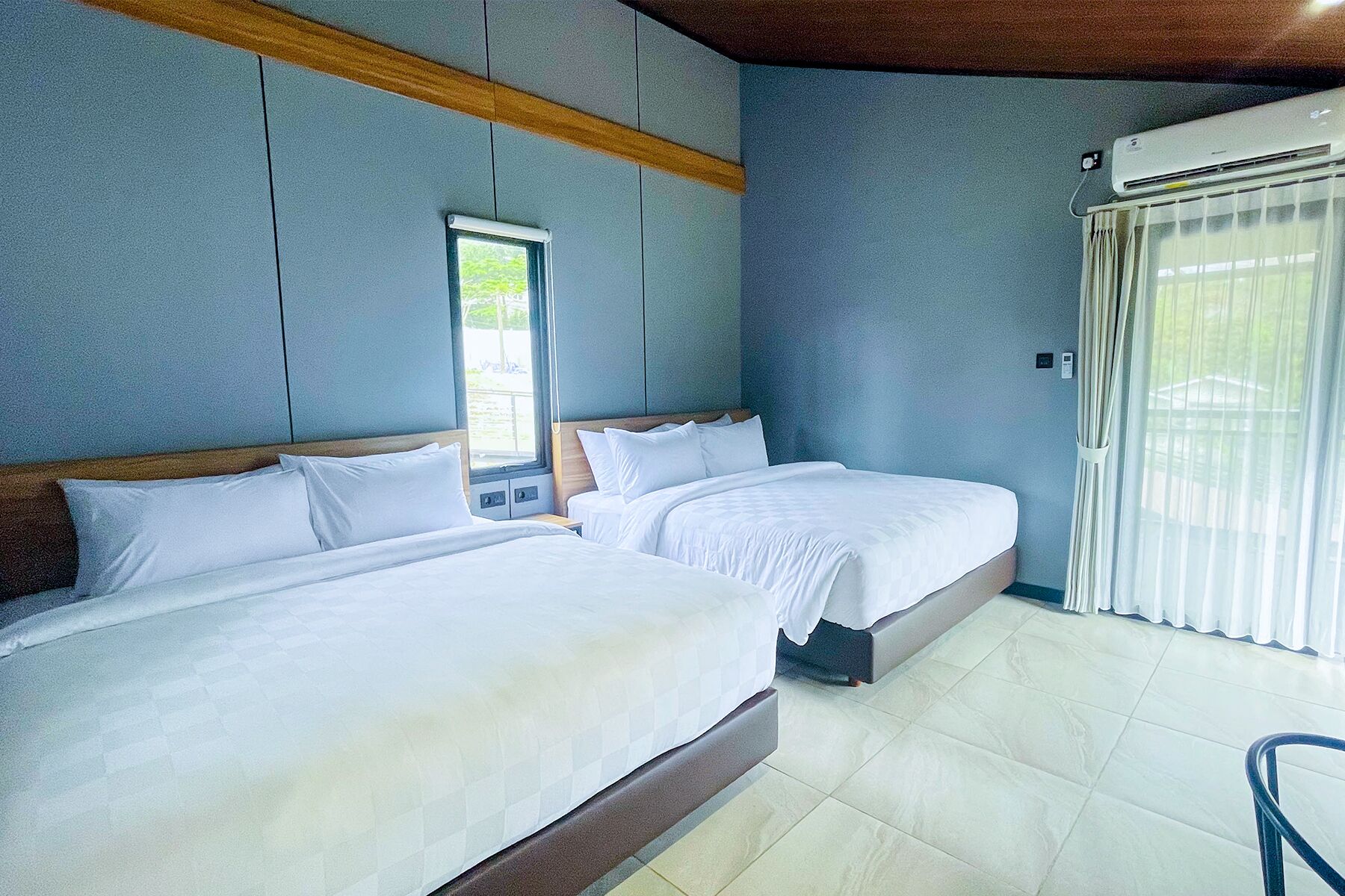 A hotel room with two beds, a window, an air conditioning unit, and a view of greenery outside.