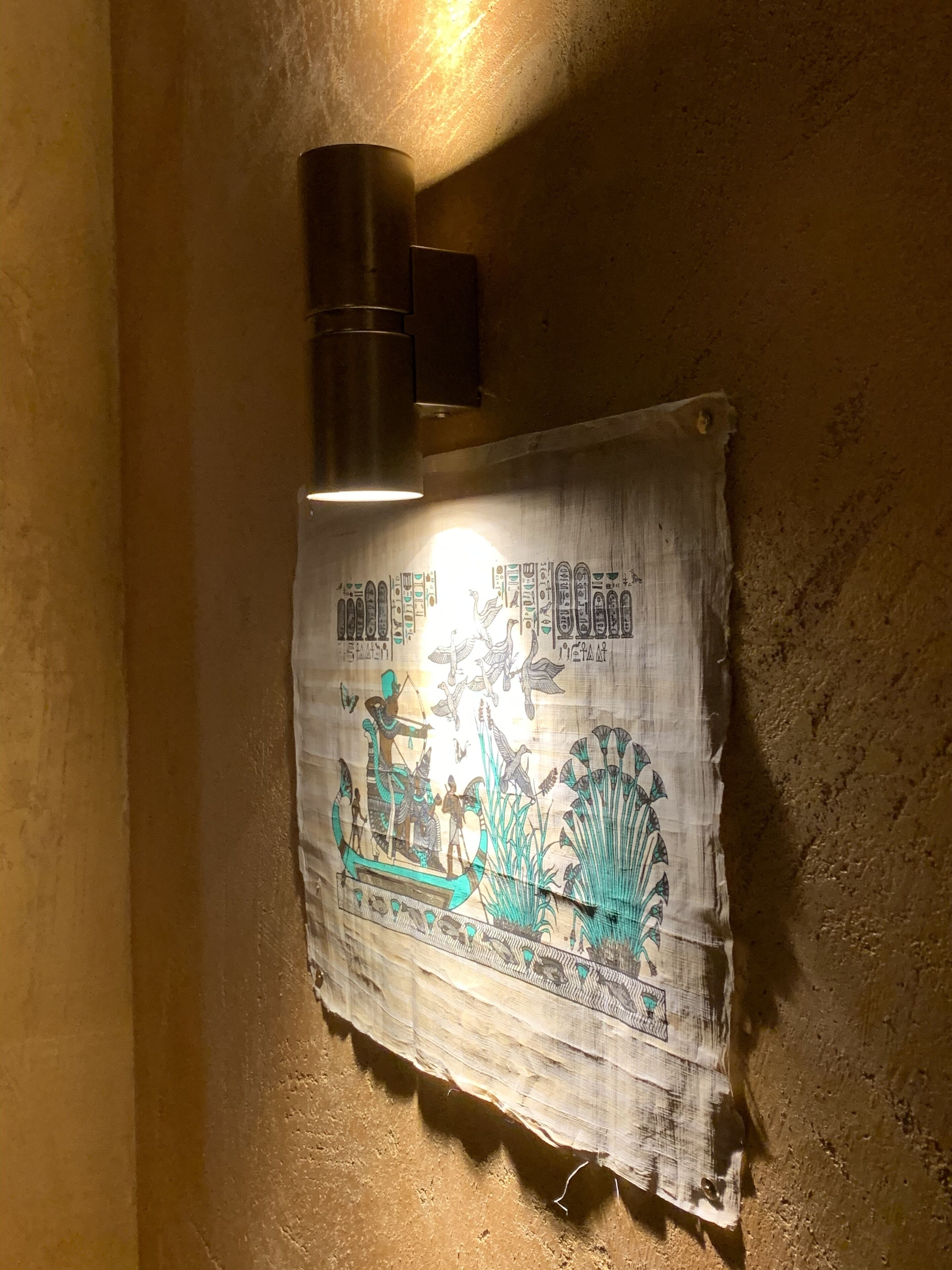 Pared decorada con un elemento que presenta jeroglíficos y figuras del antiguo Egipto, iluminada por una lámpara.