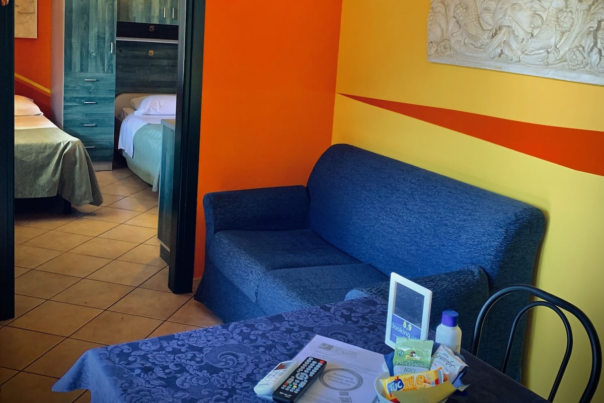 A room with a blue sofa, a TV mounted on the wall, and a table with a remote control, a bowl, and some items.