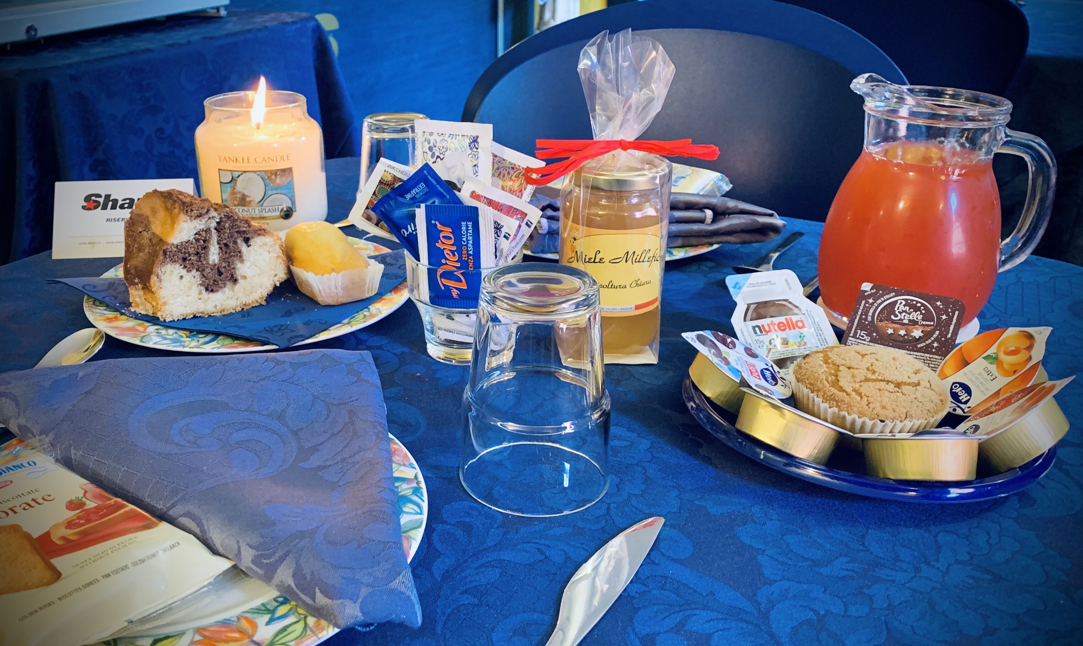 A table set with a variety of food items, including a cake, muffins, and a pitcher of juice, along with a candle and some packaged snacks.