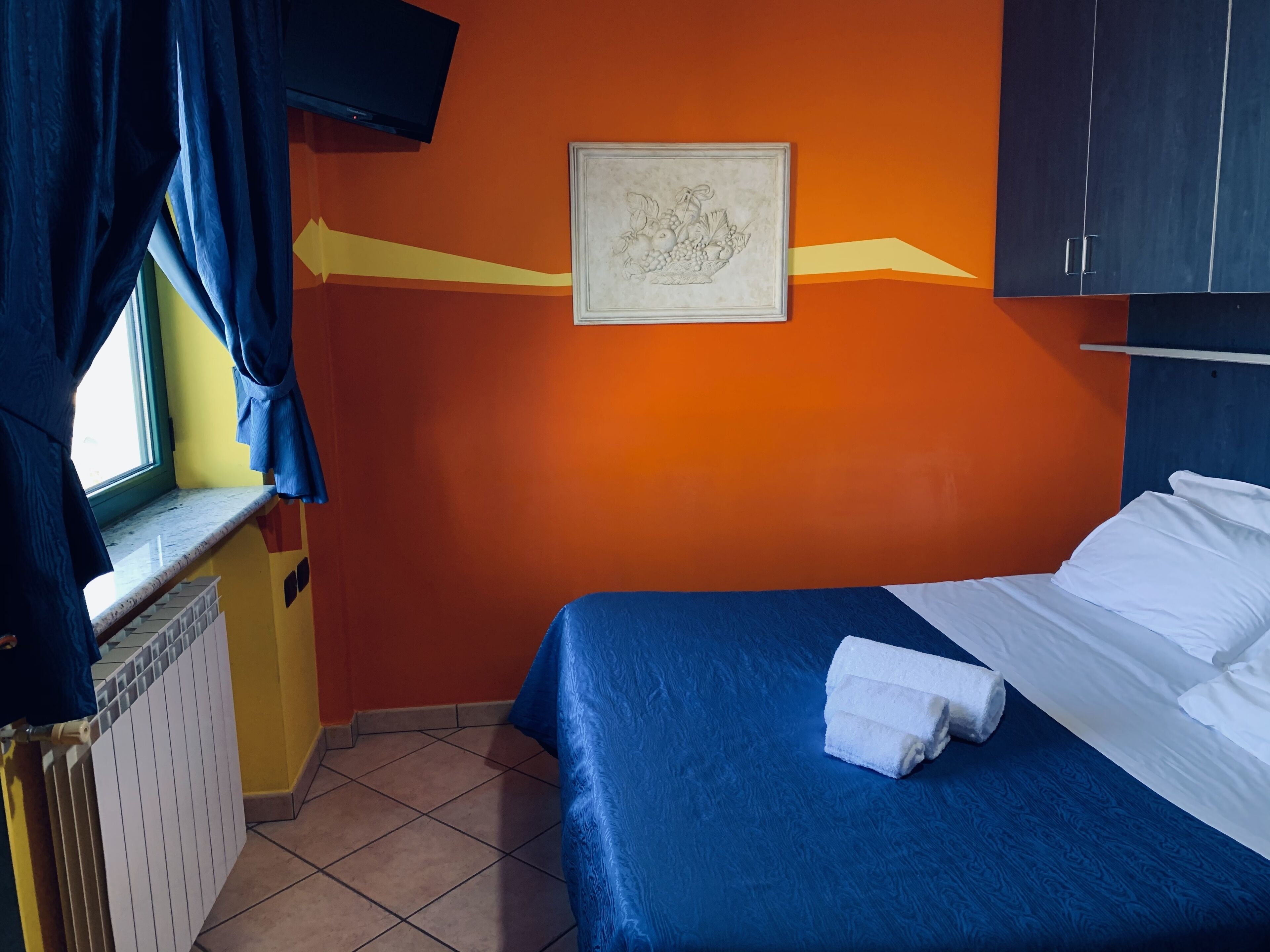 Een kamer met een bed, een raam met gordijnen, een radiator en een aan de muur bevestigde televisie.