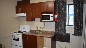 A compact kitchen with wooden cabinets, a white stove, a microwave, and a small window with floral curtains. - Hope Inn and Suites (Hope)