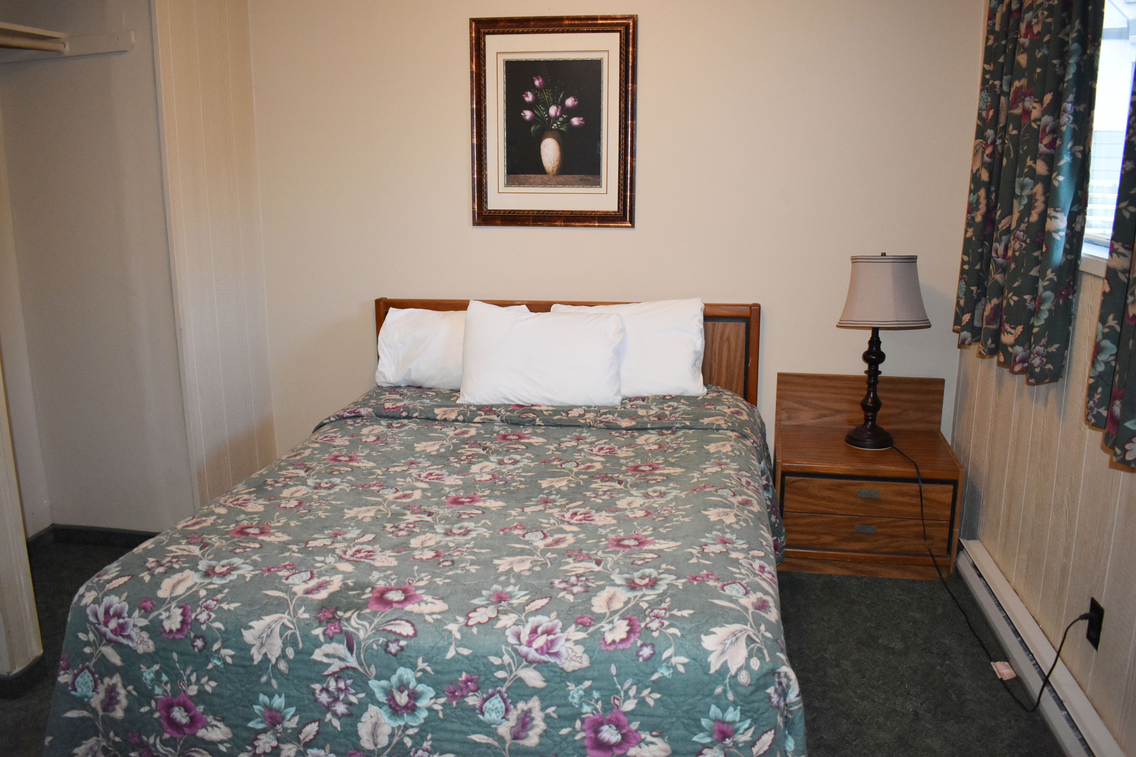 A bedroom with a floral bedspread, a wooden headboard, a bedside table with a lamp, and a framed picture on the wall.
