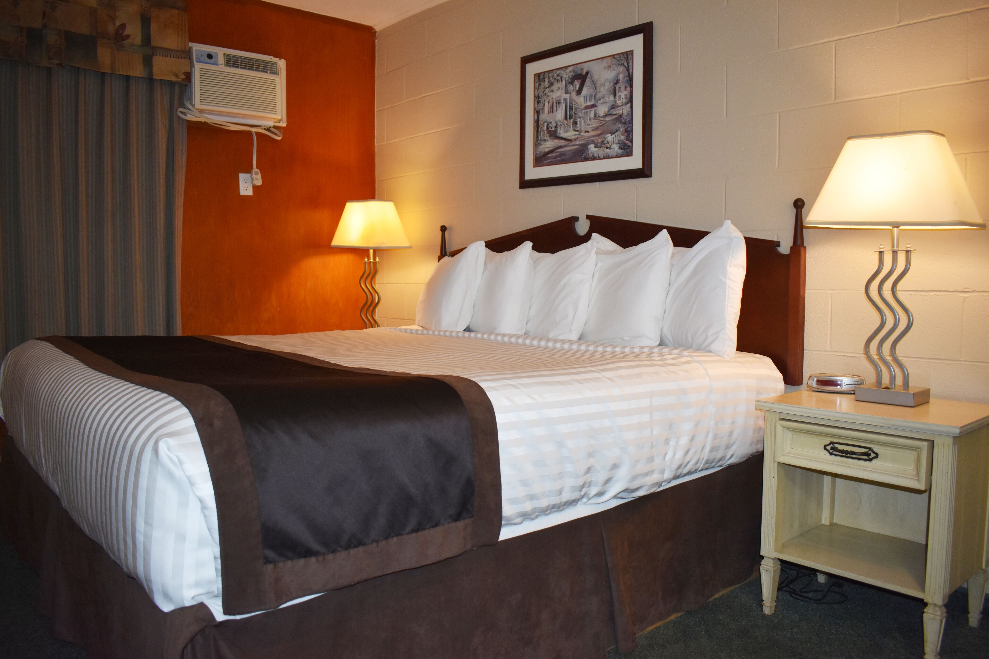 A hotel room with a bed, two bedside lamps, a nightstand, a wall-mounted air conditioner, and a framed picture.