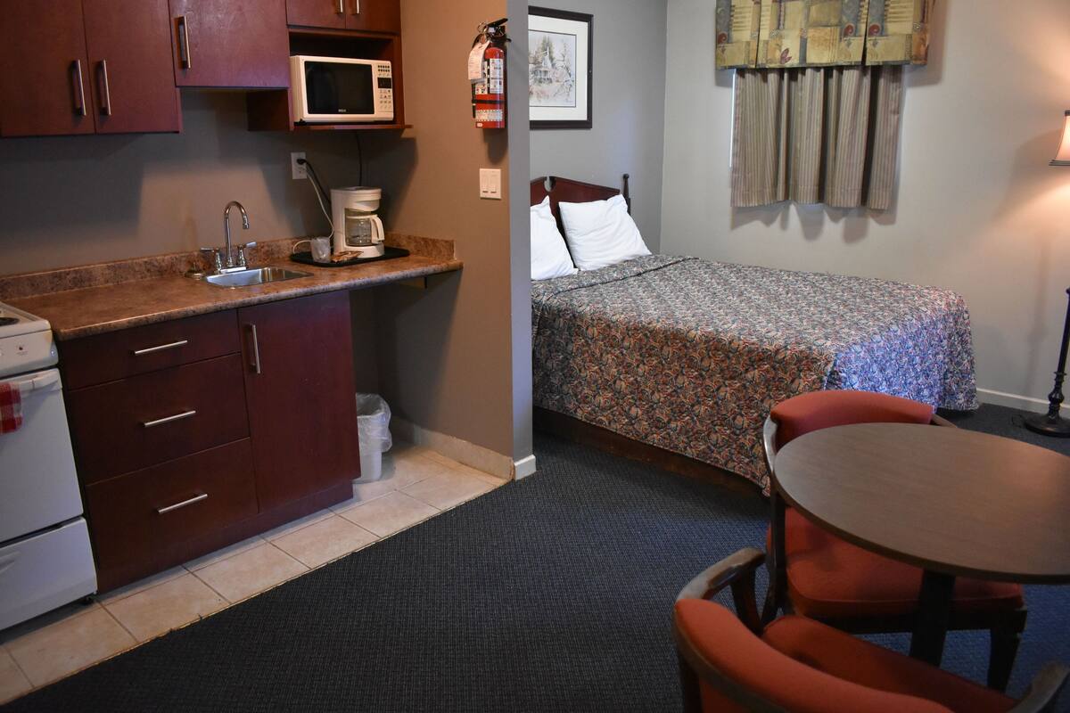 Una habitación de hotel compacta con kitchenette, una cama individual, una mesita y una silla.