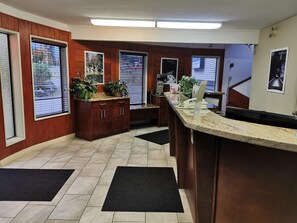 A reception area with a wooden counter, a telephone, and a computer. There are potted plants on the counter and a door with a poster on the wall.