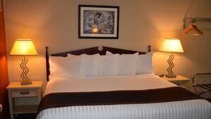 A neatly made bed with white linens and a dark headboard in a hotel room. There are two bedside lamps on either side of the bed, and a framed picture hangs on the wall above. - Hope Inn and Suites (Hope)