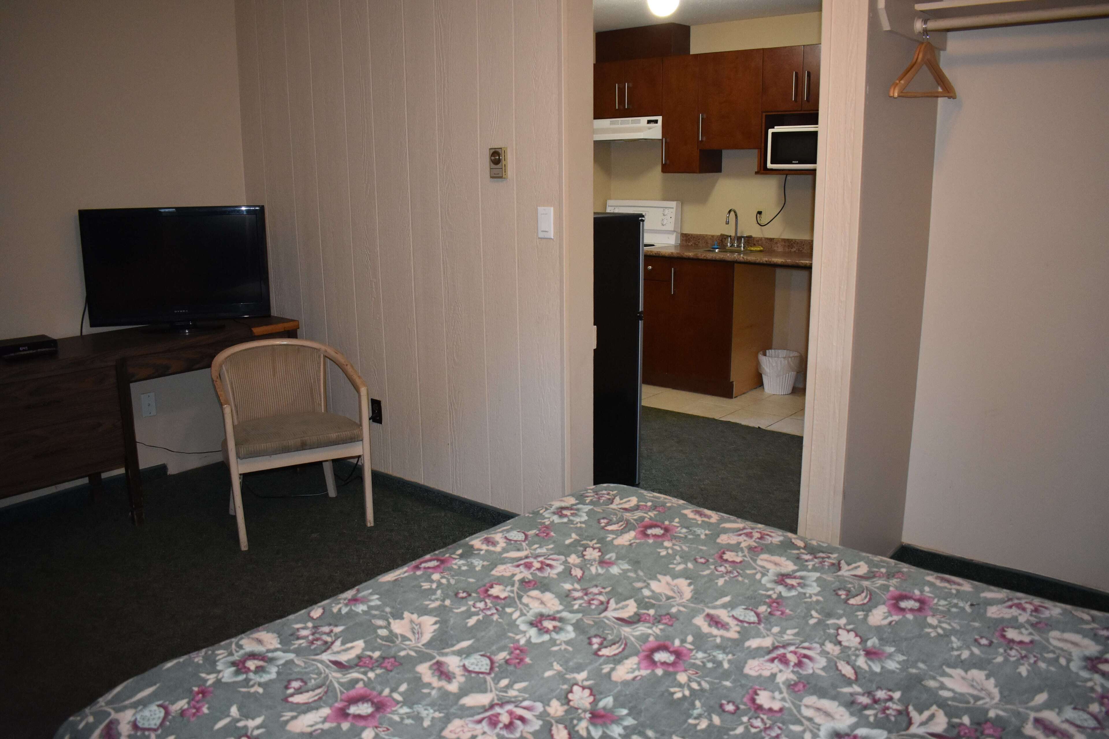 A hotel room with a bed, a desk with a television, a chair, a kitchenette with a microwave, and a refrigerator.