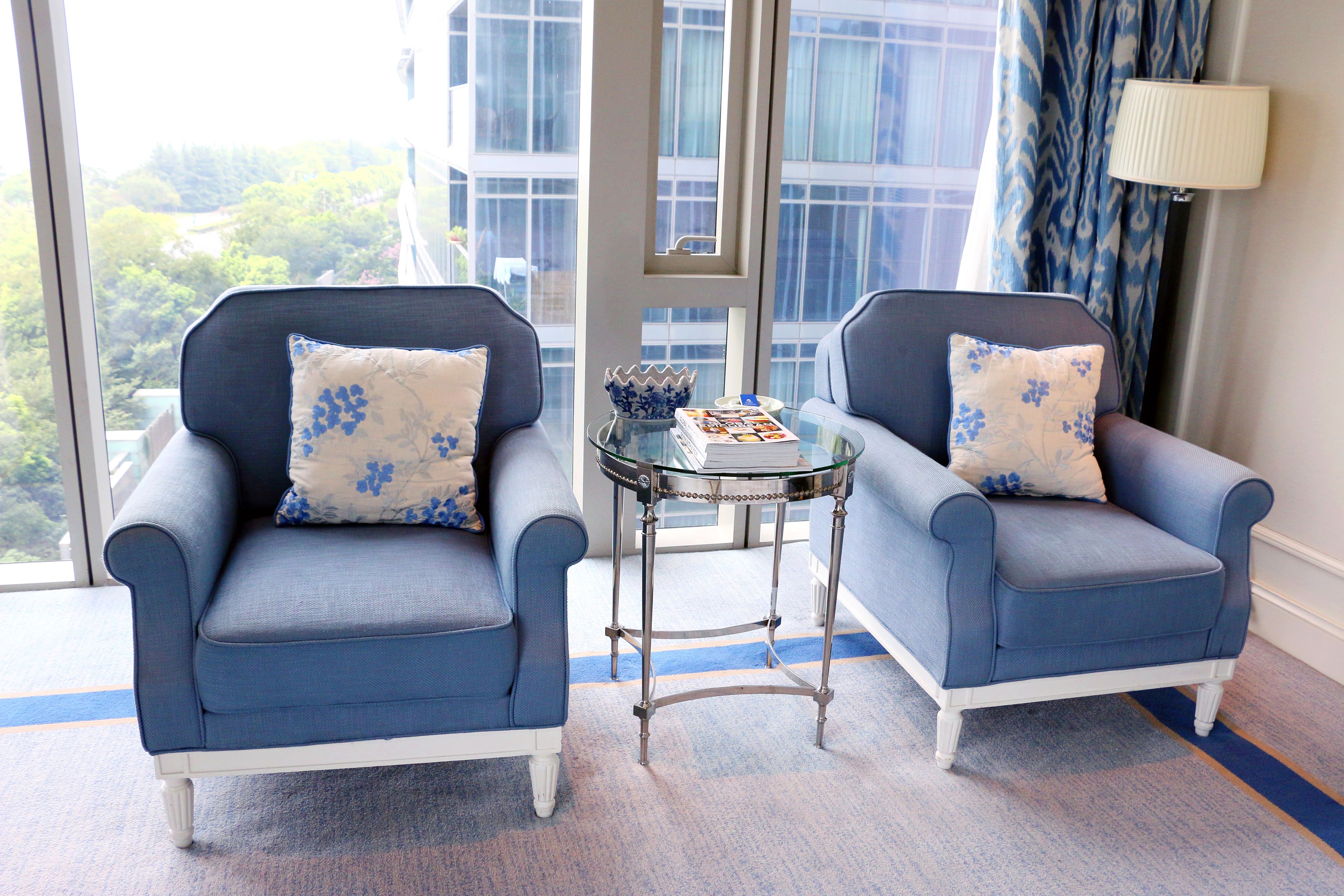 Two armchairs with floral cushions, a glass table with a bowl and book, and a view of a building and trees through large windows.