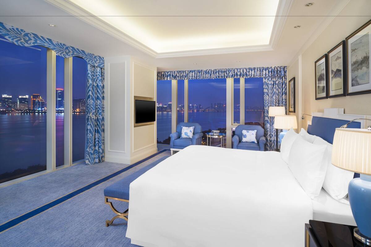 A hotel room with a large bed, two armchairs, a TV, and a view of the cityscape at night.