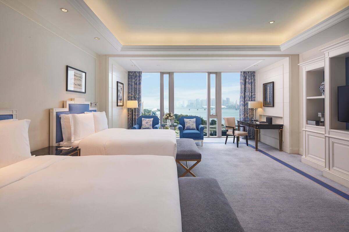 A hotel room with two beds, a desk, and a view of a cityscape.