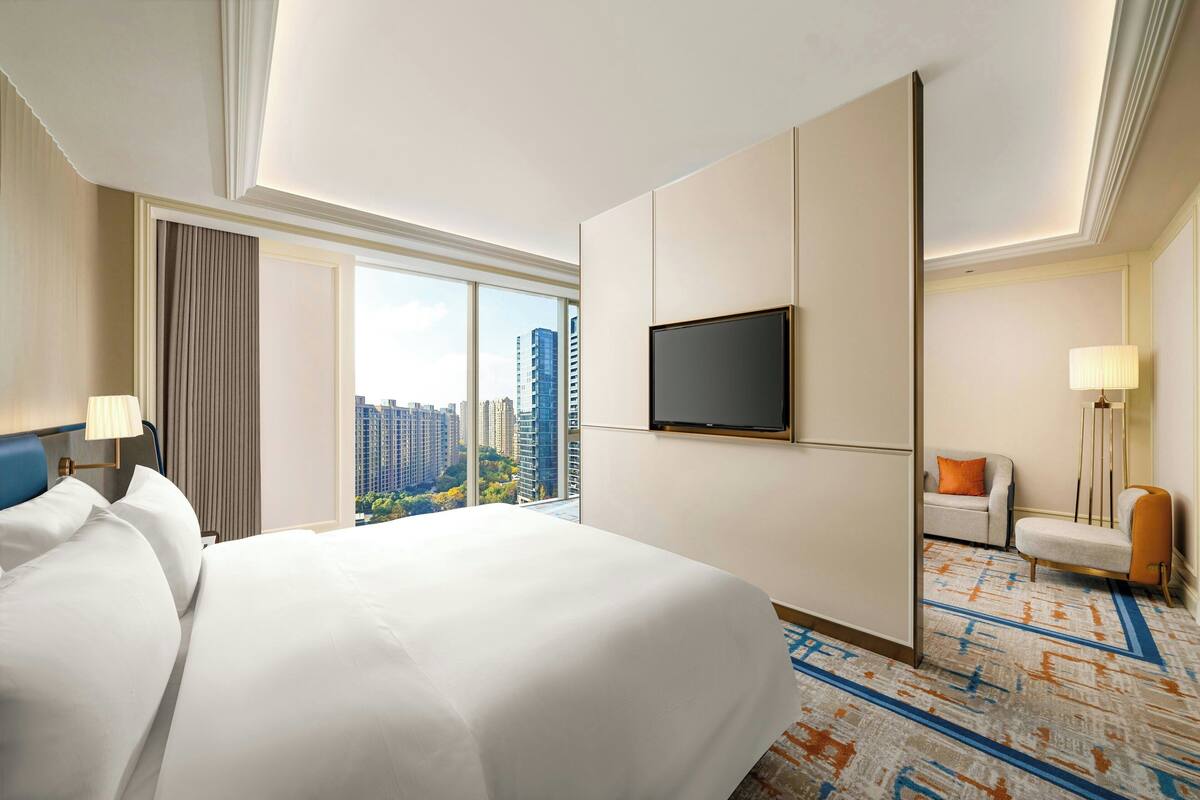 A hotel room with a large bed, a TV mounted on the wall, a sofa, and a view of the cityscape through the window.