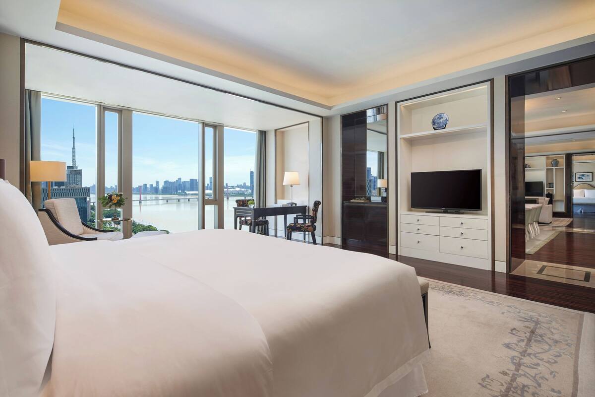 A modern hotel room with a large bed, a desk with a chair, a television, and a view of the city skyline.